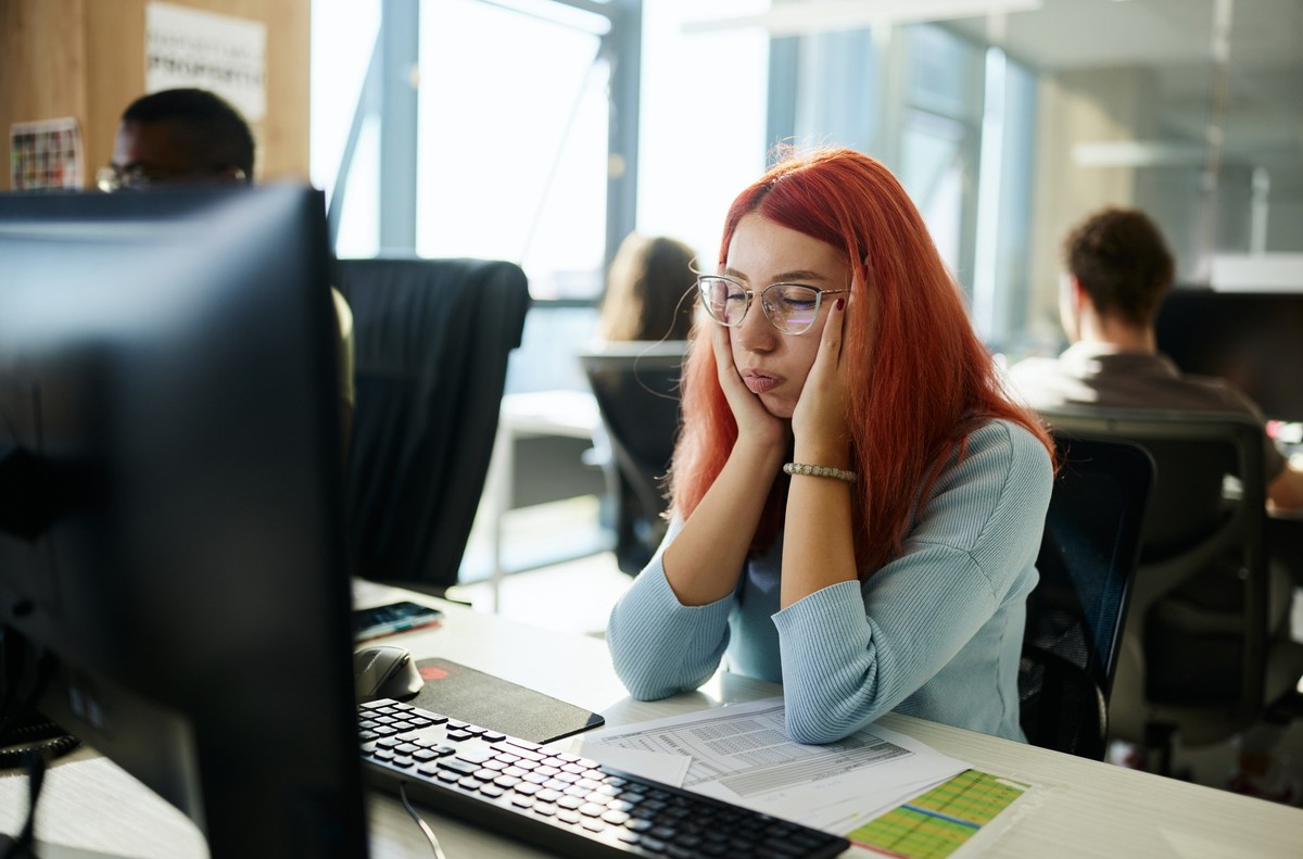 Young redhead female designer feeling exhausted while working on desktop computer in the office.