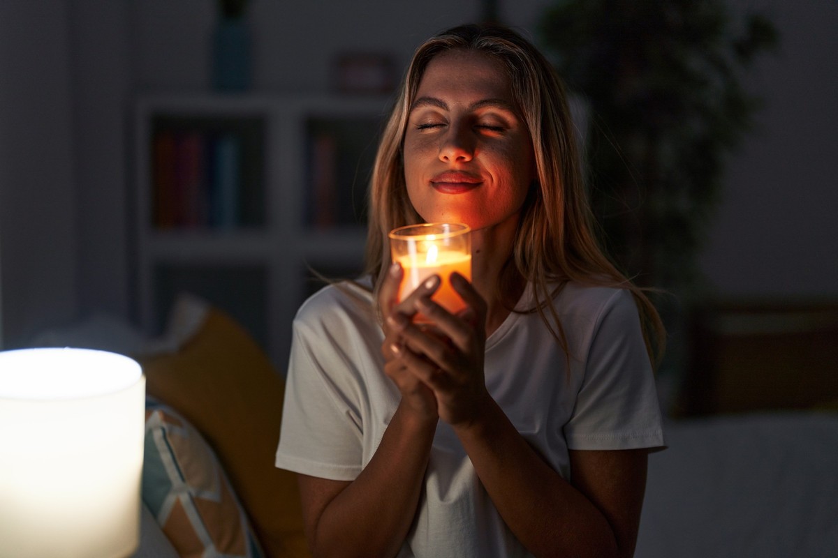 Young blonde woman sitting on bed smelling aromatic candle at bedroom
