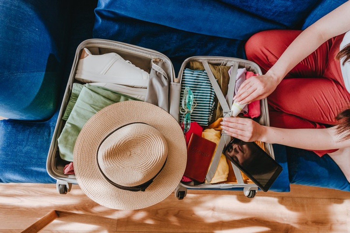 Top view of beautiful young woman packing personal belongings in a suitcase for travelling looking at list at tablet sitting on the sofa in the living room.