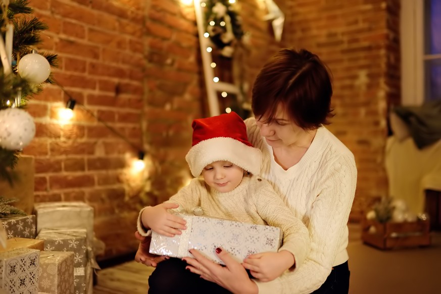 Cute little boy wearing Santa hat and his mother or grandmother opening a Christmas gift. Portrait of happy family on Christmas eve. Cozy decorated living room