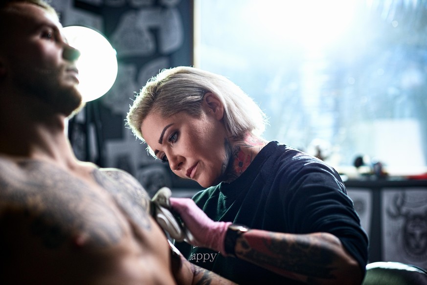 Woman tattooing man in tattoo parlour, looking and concentrating, wearing protective gloves, skill, focus, control