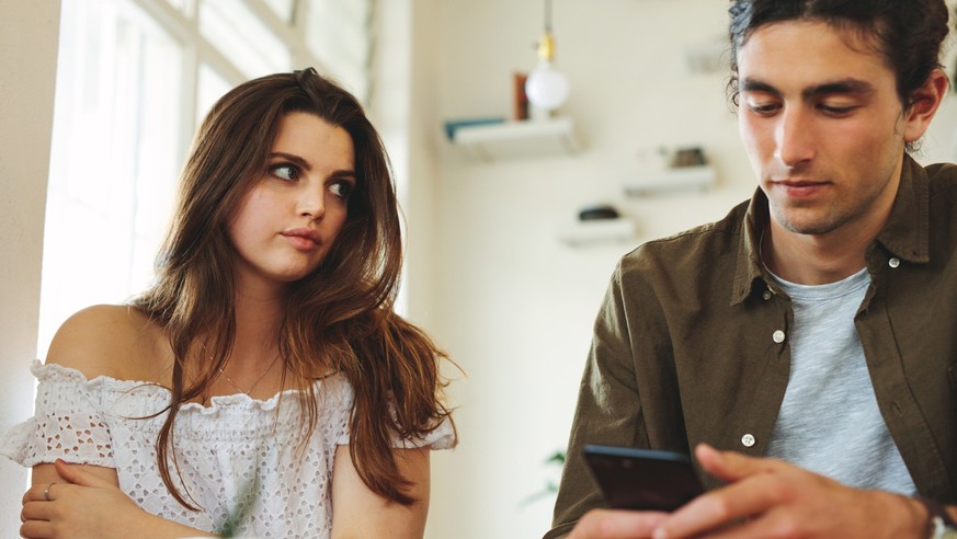 Woman looking unhappy while her man paying no attention to her and busy using his mobile phone. Sulking woman sitting next to man reading text messages during a date.