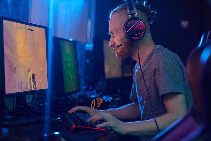 Young bearded man in headphones looking at computer monitor and playing in computer game alone in dark room