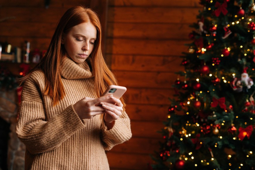 Sad young woman typing message on mobile phone while self-isolating during the winter holidays at room with festive interior. Concept of celebrating Christmas and New year at alone while lockdown.