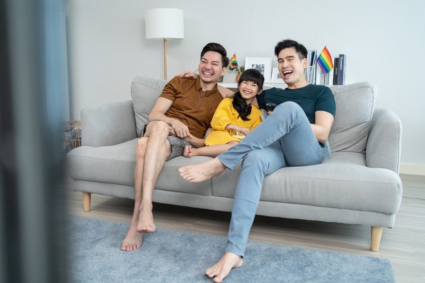 Asian male gay family with young daughter watch movie in living room. Little adorable girl child sit on sofa with attractive romantic male lgbt couple enjoy comedy show on TV and eat popcorn together.