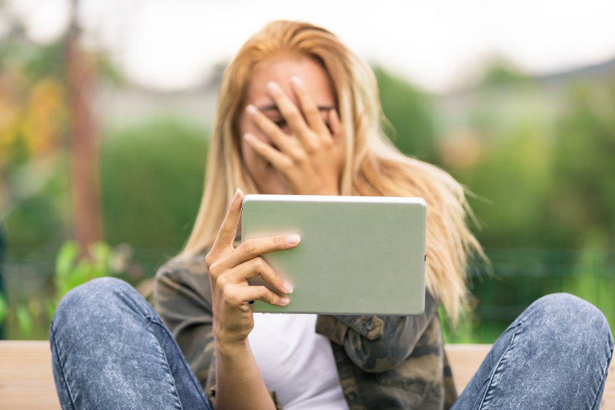 young woman hiding his smiling face because she's embarassed by some porn video or another forbidden thing she saw on the internet using her tablet outdoors