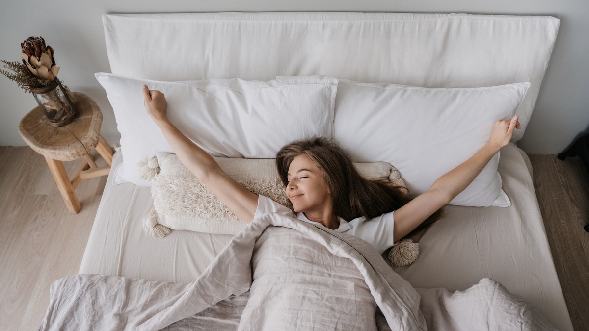 Young long haired Italian girl laying on bed eyes closed awakening at Sunday morning smiles satisfied by healthy sleep. Top view on young woman spreading hands, stretching. Cozy home, happy people.