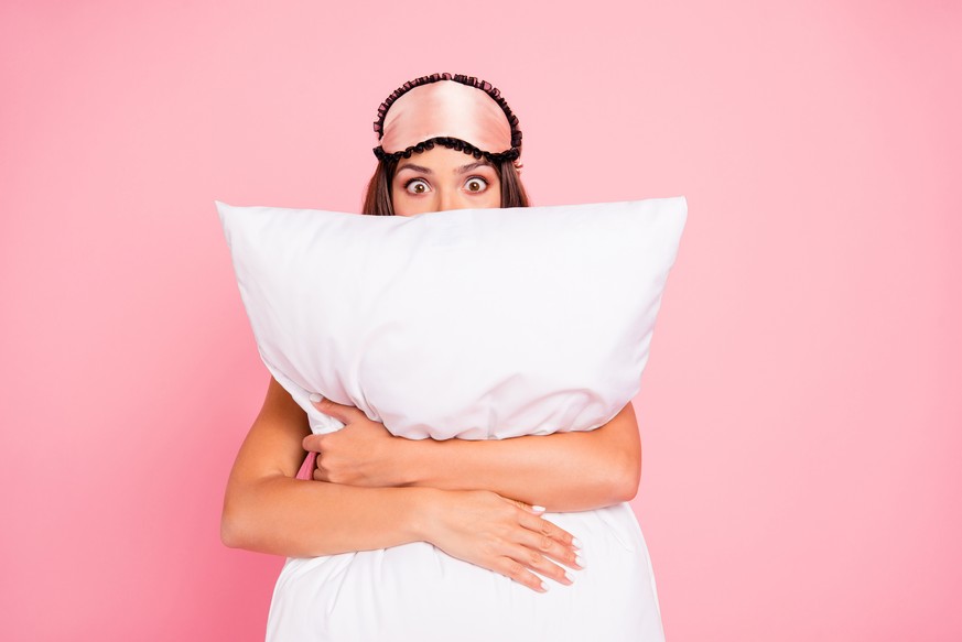 Young gorgeous shocked lady wearing eye mask, hugging pillow, hiding. Isolated over pink pastel background