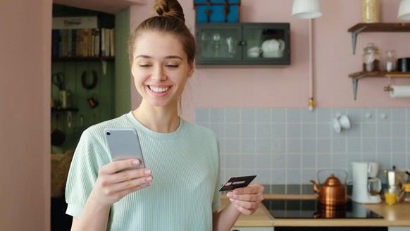 Indoor shot of young European girl pictured in her flat holding smartphone in one hand and credit card in another, processing online payments via banking application to purchase goods with content