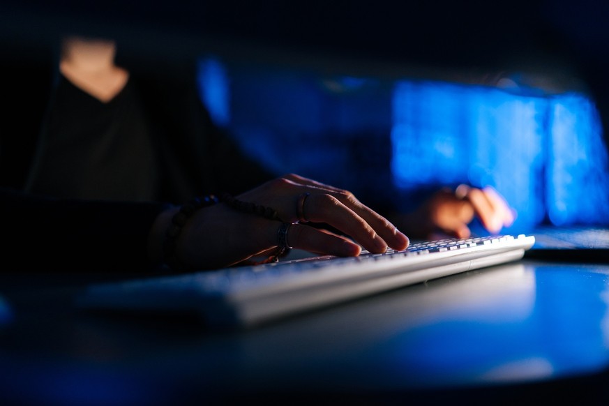 Close-up hands of unrecognizable hacker man working typing on keyboard laptop computer sitting at desk in dark room with blue neon lights. Concept of cyber attack, virus, malware, illegally.