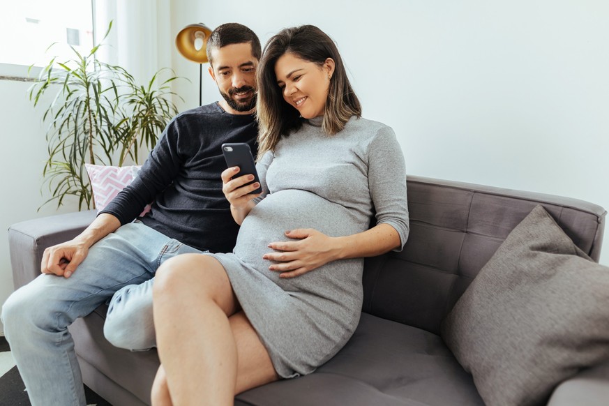 Adult couple expecting baby spending time together at home. Couple using smartphone.