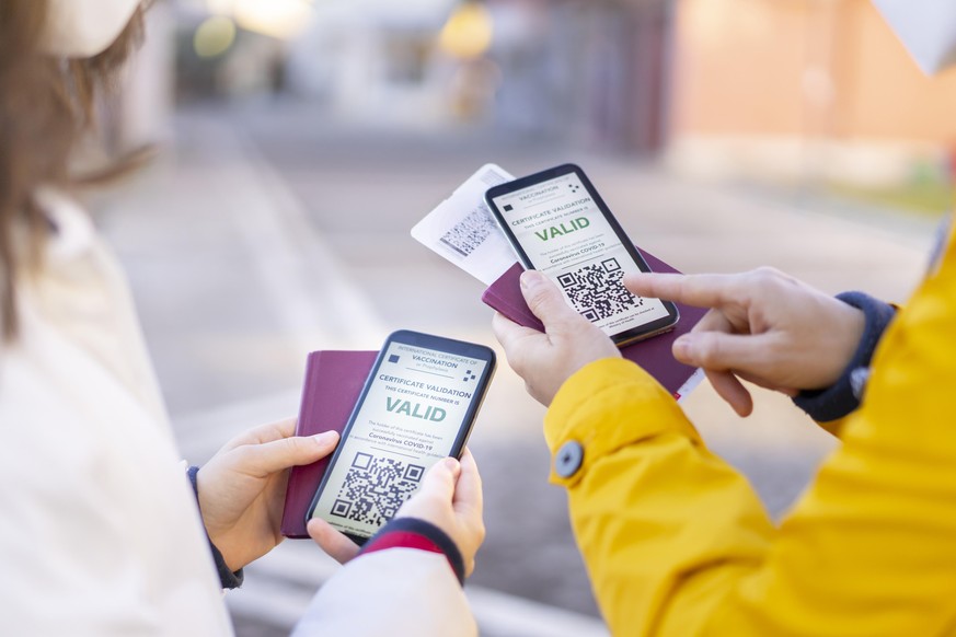 Friends checking their digital international certificates of COVID-19 Vaccination, standing on the street, wearing a FFP2 Mask, holding boarding passes and pasports