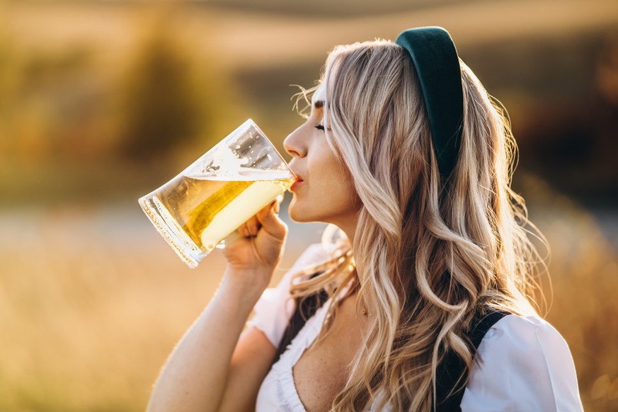 Pretty happy blonde in dirndl, traditional festival dress, drinking beer outdoors in the field with blurred background. Beer Fest, St. PatrickÃ¢Â€Â™s day, international beer day concept.