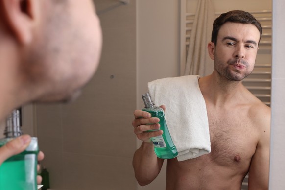 A handsome shirtless man in his forties is using mouth wash in the bathroom. He looks at himself in the mirror while doing it.