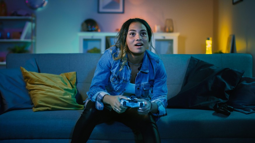 Beautiful Excited Young Black Gamer Girl Sitting on a Couch and Playing Video Games on a Console. She Plays with a Wireless Controller. Cozy Room is Lit with Warm and Neon Light.