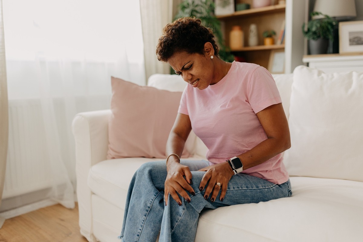 A woman in casual attire is seated on a white sofa, touching her knee in apparent discomfort, in a modern, well-decorated living room space filled with natural light.