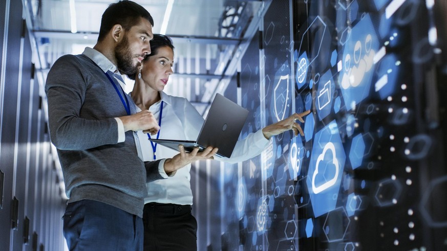 Male IT Specialist Holds Laptop and Discusses Work with Female Server Technician. They&#039;re Standing in Data Center, Rack Server Cabinet with Cloud Server Icon and Visualization.