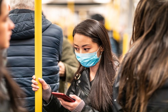 Chinese woman wearing face mask in the train to protect from virus - young asian woman looking worried and holding a smartphone with many people all around - health and travel concepts