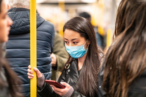 Chinese woman wearing face mask in the train to protect from virus - young asian woman looking worried and holding a smartphone with many people all around - health and travel concepts