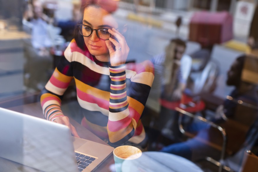 Beautiful Young Woman with Long Black Hair and Glasses Using a Laptop while Drinking Coffee or Tea at Cafe.The reflection from the glass Windows