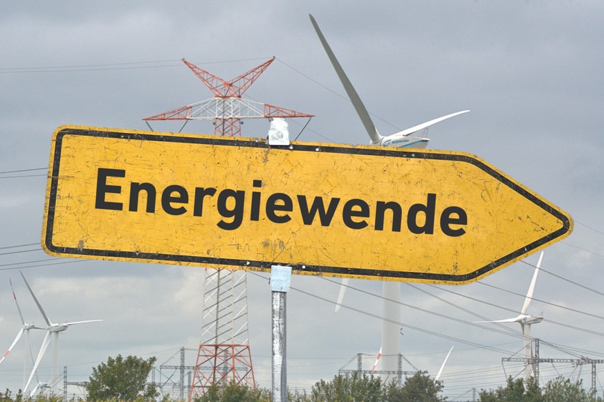 [M] Ein gelbes, zerkratztes Hinweisschild mit der Aufschrift &quot;Energiewende&quot; vor einigen Windkraftanlagen und einem im Aufbau befindlichen Freileitungsmast. Composing als Symbolbild in Bezug  ...