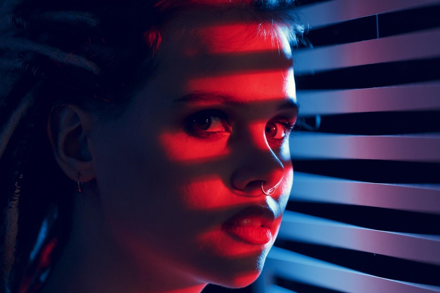 Portrait of a young woman in neon light, looking through the blinds. A trendy portrait with colored lighting and striped shadows.