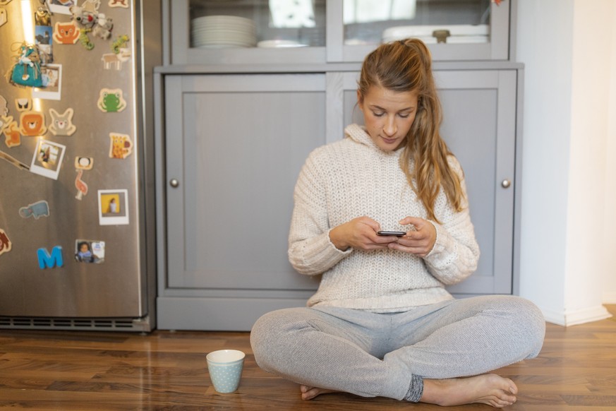 Young woman sitting on the floor in kitchen at home using cell phone model released Symbolfoto property released PUBLICATIONxINxGERxSUIxAUTxHUNxONLY GUSF02474