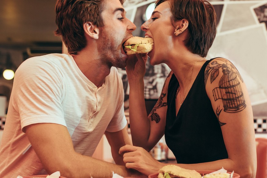 Romantic couple eating a burger together at a restaurant looking at each other. Couple dining at a restaurant biting a burger together. symbiose