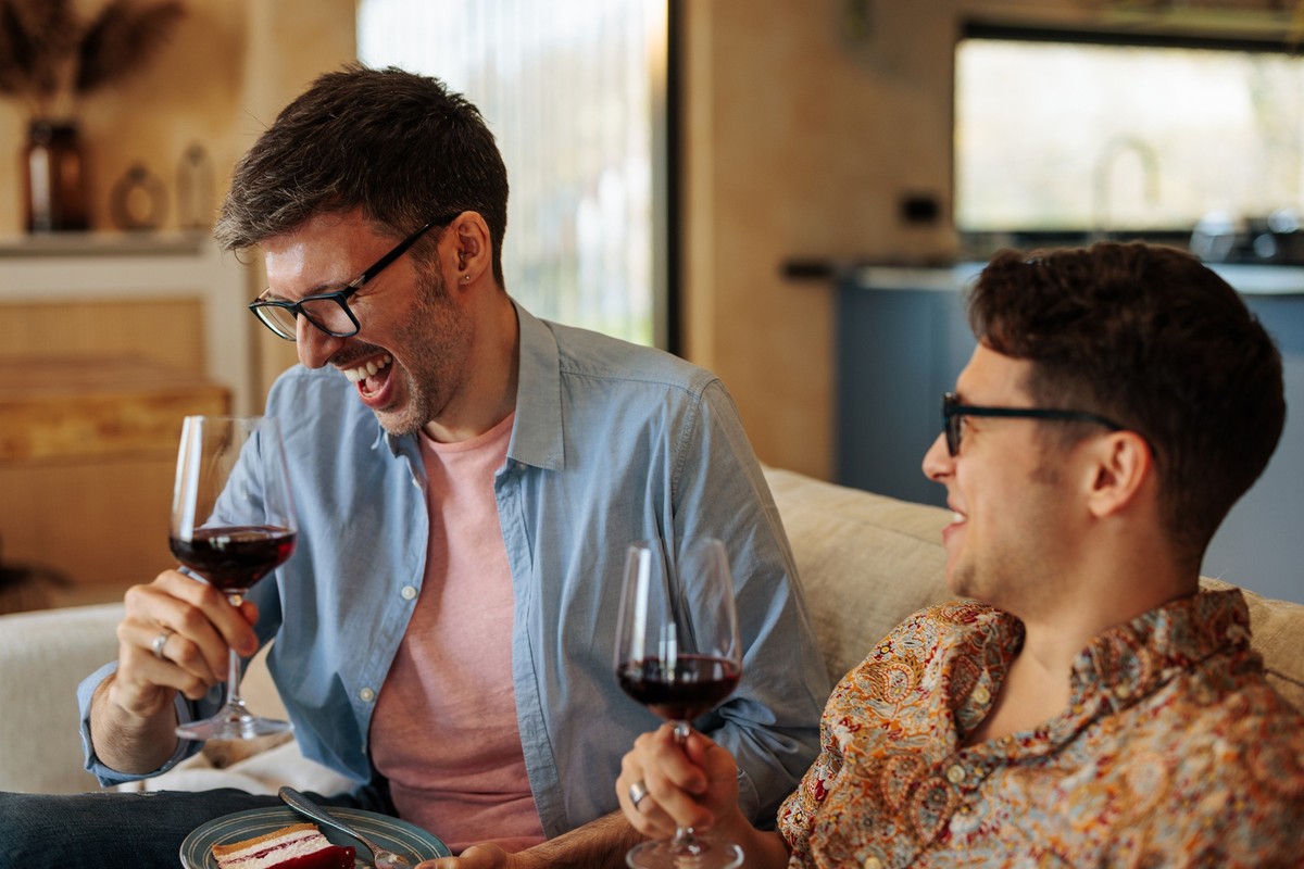 A cheerful gay couple is at home, drinking wine on the sofa in the living room and laughing.