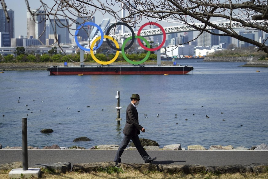 Ein Mann spaziert vor der Stadtkulisse der im Wasser schwimmenden Olympischen Ringe in Tokyo.