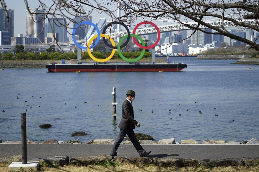 18.03.2021, Japan, Tokyo: Ein Mann spaziert vor der Stadtkulisse der im Wasser schwimmenden Olympischen Ringe im Stadtteil Odaiba. Foto: Eugene Hoshiko/AP/dpa +++ dpa-Bildfunk +++