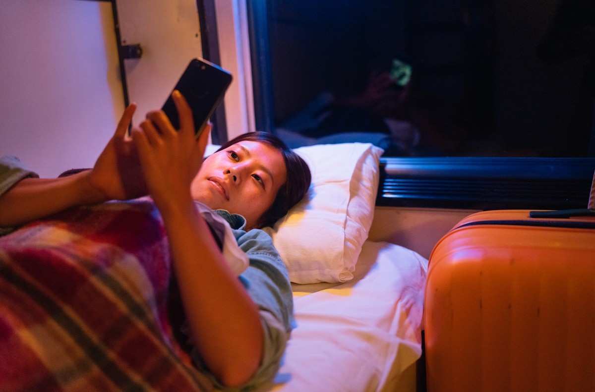 A young female traveller is lying down in a bed in a train compartment comfortably and using a smart phone while traveling at night.
