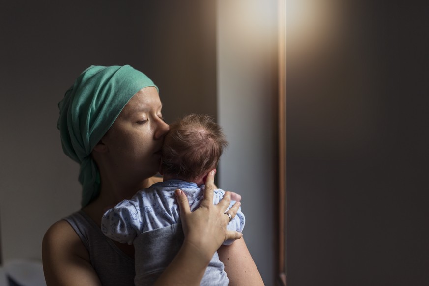 Caucasian Pensive Woman in headscarf, fighting breast cancer while holding her newborn baby relaxing in cancer treatment hospital, patient standing next to hospital window. Mother and baby son. Sleepy ...