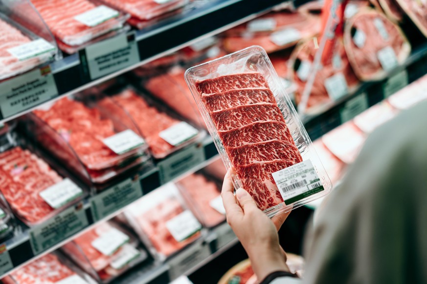 Over the shoulder view of young Asian woman shopping in a supermarket. She is choosing meat and holding a packet of organic beef in front of the refrigerated section