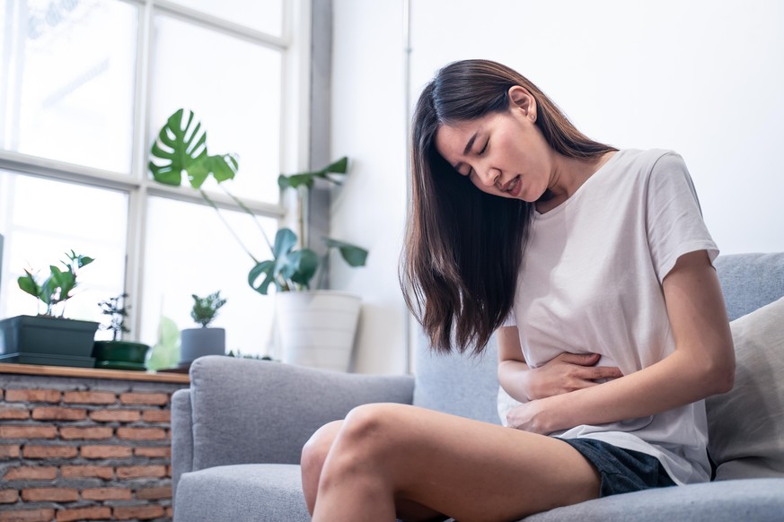 Asian young woman sick with food poisoning. She sit on sofa at home. Female have stomach ache and curls through spasms. Girl feel so much painful, sad and lonely. Healthcare food and medical concept.