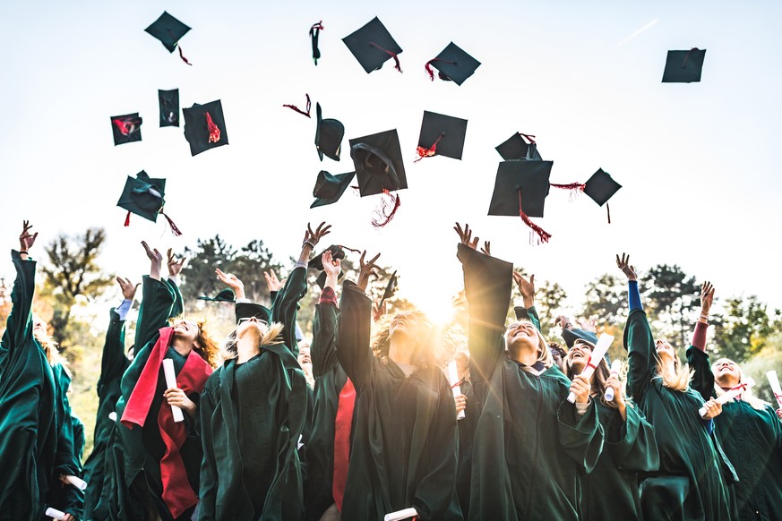 Large group of happy college students celebrating their graduation day outdoors while throwing their caps up in the air.