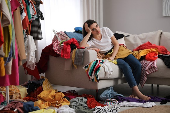 Upset young woman with lots of clothes on sofa in room. Fast fashion