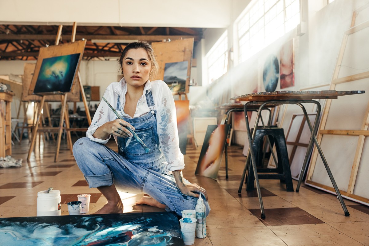 Inventive female painter looking at the camera in her art studio. Skilful artist making a blue painting for her new art project. Creative young woman working on the floor in her atelier.