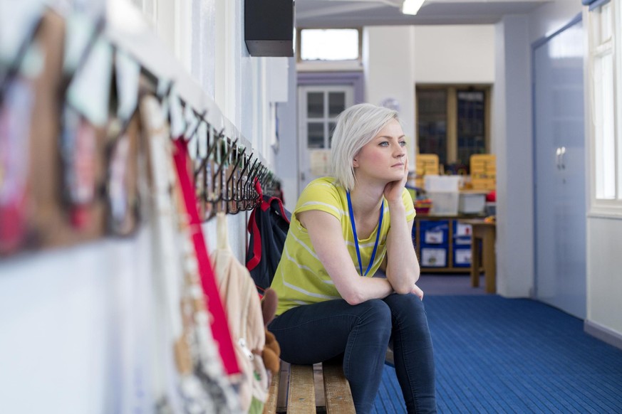 Young, female teacher sitting on a bench in the cloakroom of school. She is looking pensively out of the window.