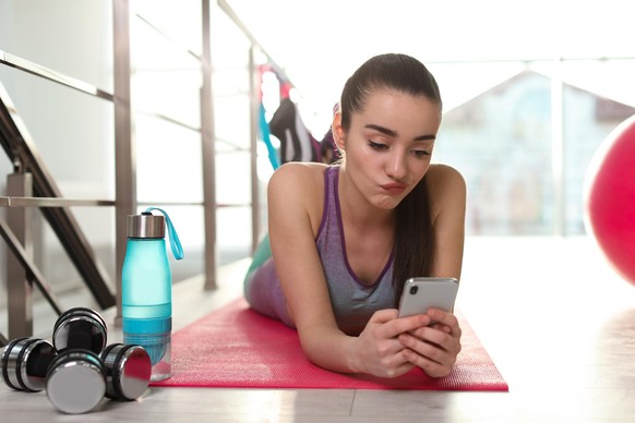 Lazy young woman with smartphone on yoga mat indoors