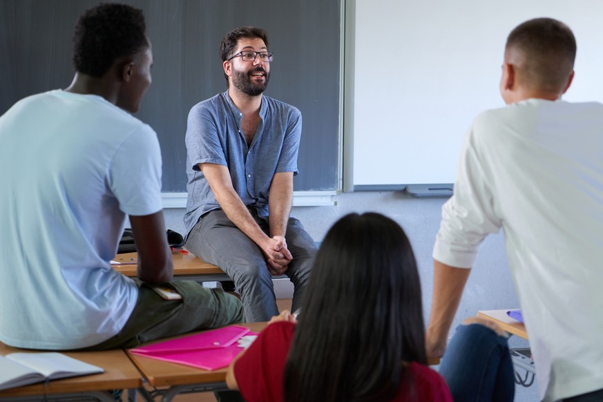 Teacher involved with students at college sitting on his desk while giving an interactive class. High quality photo