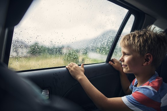 Kids aged 7 is looking out of the car window. The glass is covered by rain drops.