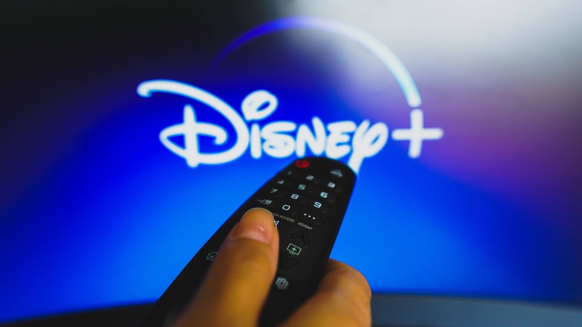 May 10, 2022, Brazil. In this photo illustration a close-up of a hand holding a TV remote control seen in front of the Disney Plus logo. May 10, 2022, Brazil. In this photo illustration a close-up of  ...
