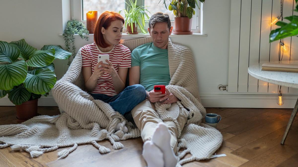 Curious woman looking at man husband smartphones screen chatting on social network. Obsessed family couple using smartphones share information between gadgets, shopping online sitting on flour at home ...