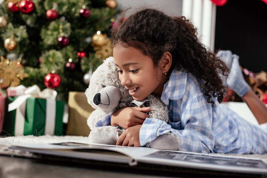 happy adorable african american child in pajamas with teddy bear looking at photo album on floor at home