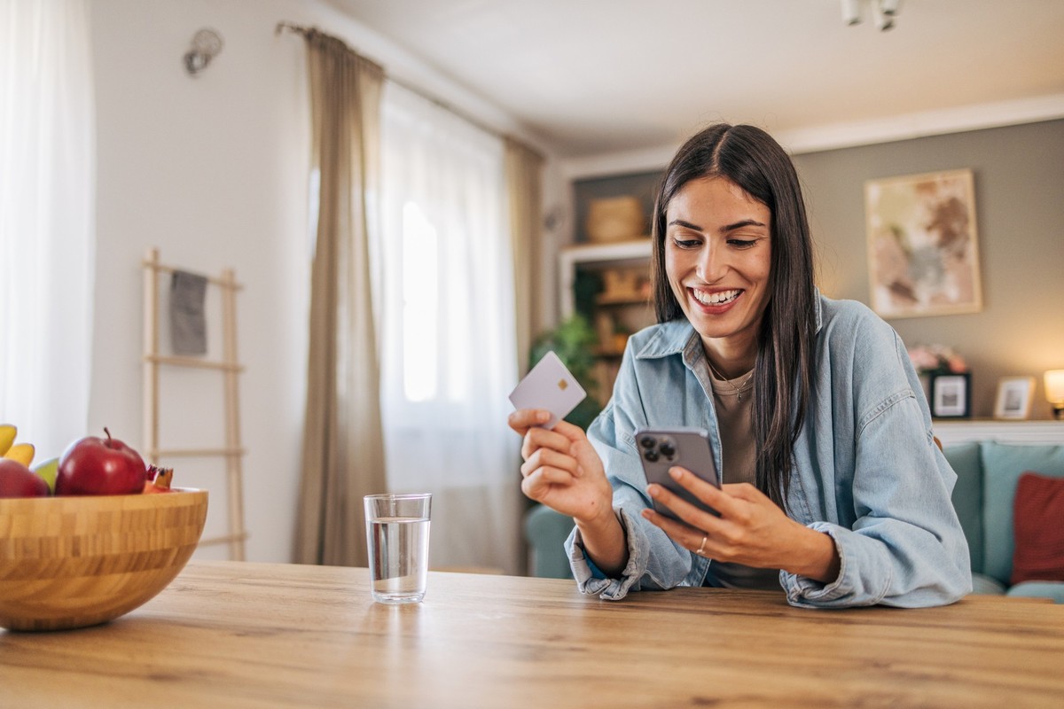 A young, cheerful woman using her smartphone to shop online and holding a credit card.