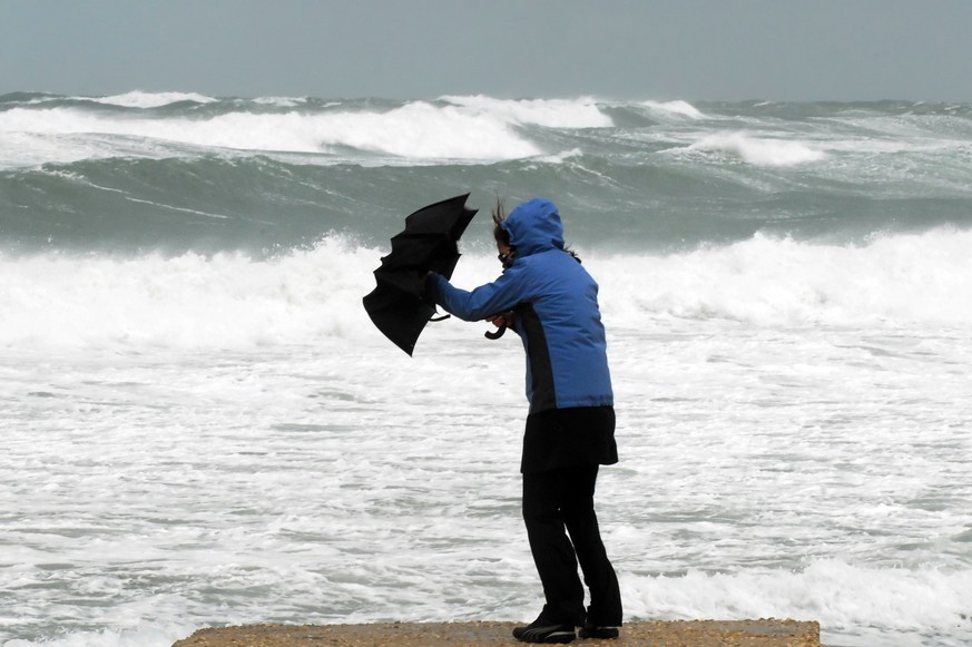 A woman in a blue raincoat holding a black umbrella struggles with the umbrella due to stormy, windy weather on a beach