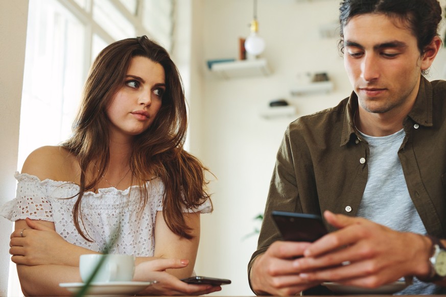 Woman looking unhappy while her man paying no attention to her and busy using his mobile phone. Sulking woman sitting next to man reading text messages during a date.