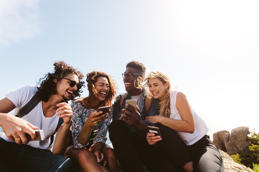 Young group of people sitting on top of mountain using smart phones and smiling. Diverse friends enjoying a day out.