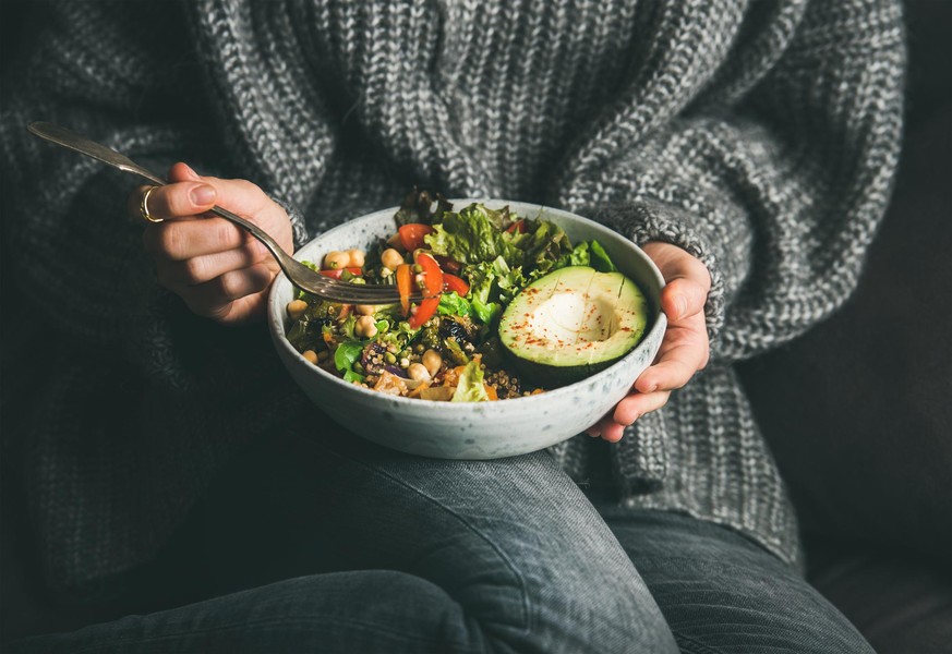 Healthy vegetarian dinner. Woman in grey jeans and sweater eating fresh salad, avocado half, grains, beans, roasted vegetables from Buddha bowl. Superfood, clean eating, dieting food concept